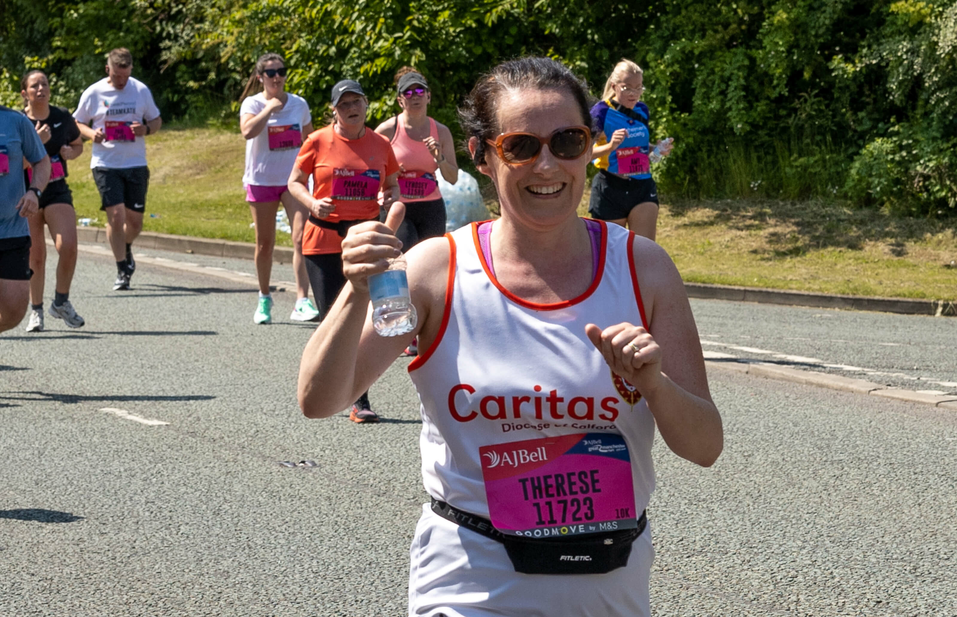 Person running in a white Caritas vest with race number. Holding a bottle of water up and smiling at camera