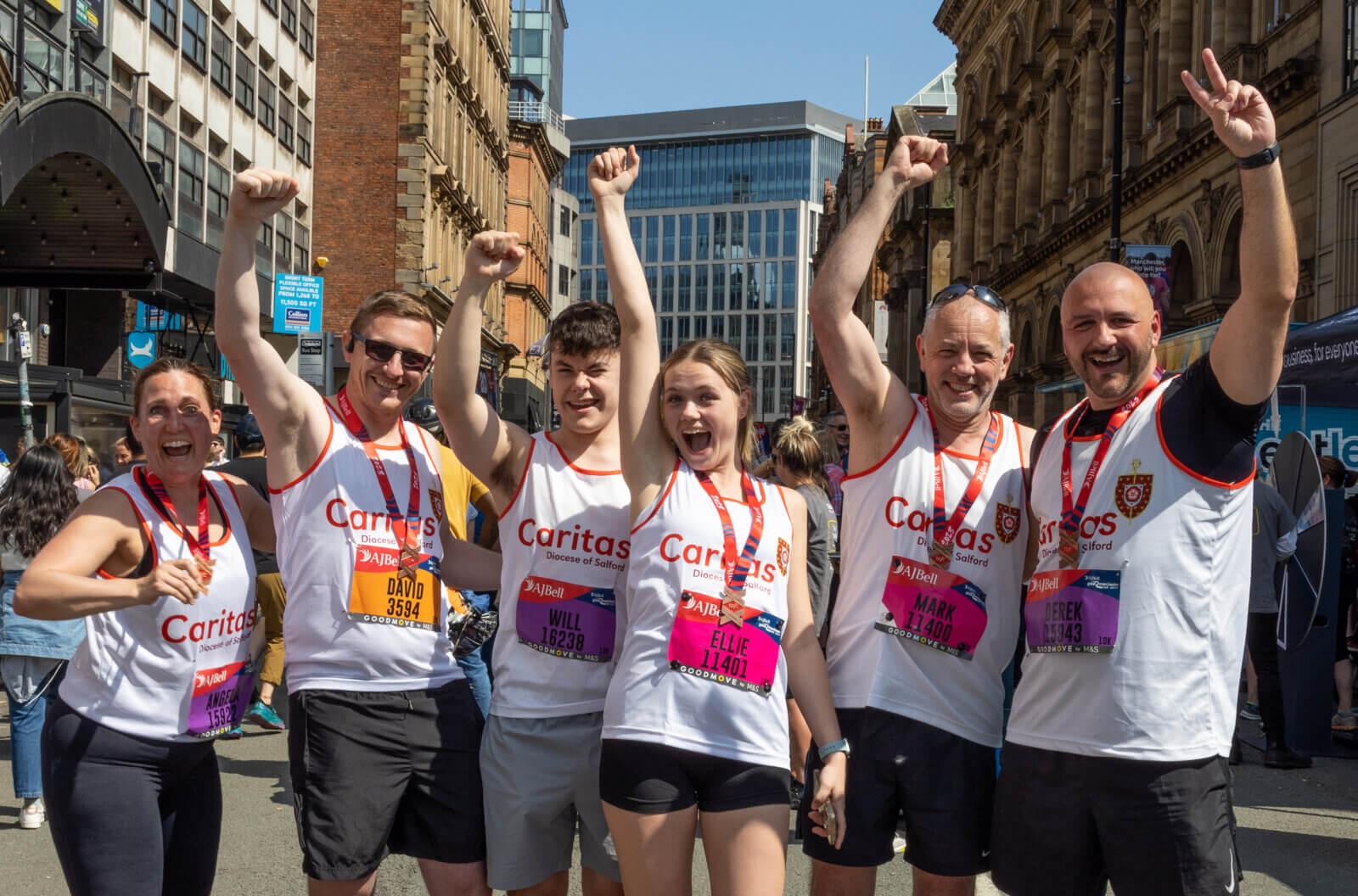 Group of people after running the Great Manchester Run. All wearing medals and Caritas branded white vests and are waving their arms in the air in celebration