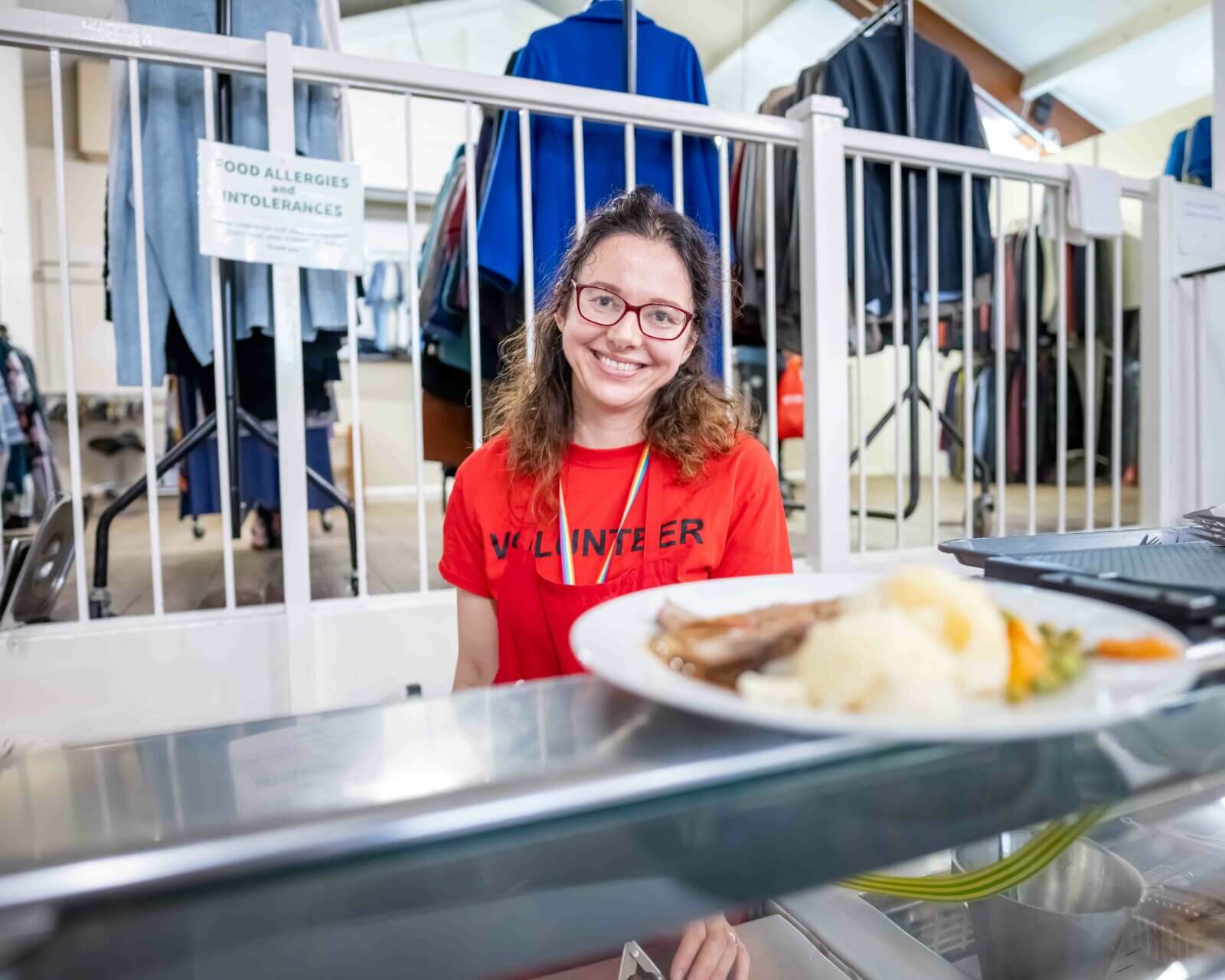 Lady in red tshirt which says volunteer on the front in black writing is smiling at camera. She is standing behind a silver food service counter with a plate of food on top