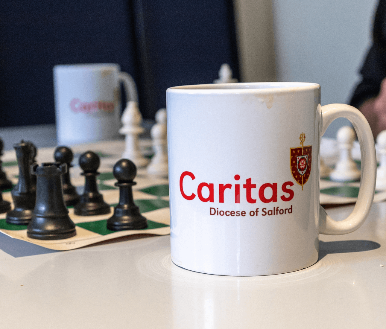 White mug with caritas logo on it. Chess board seen in background