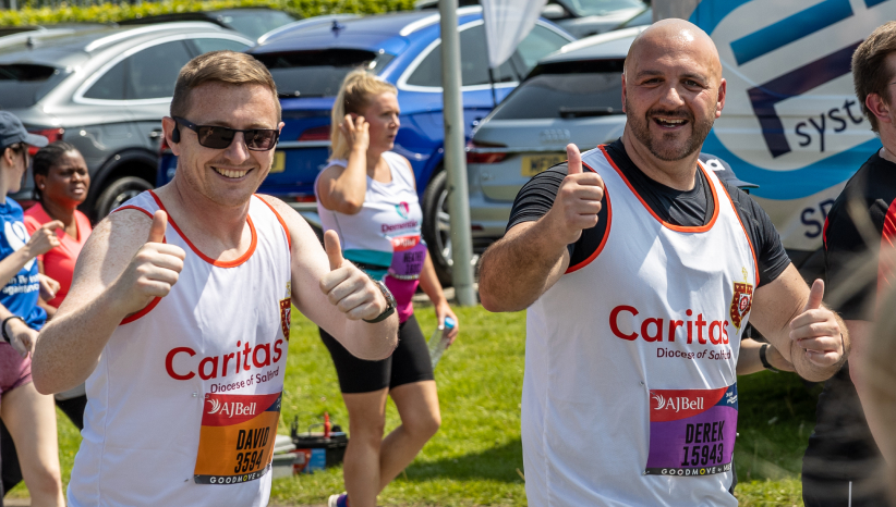 Two men in white running vests with caritas logos on front smiling at camera as they run and giving thumbs up to camera
