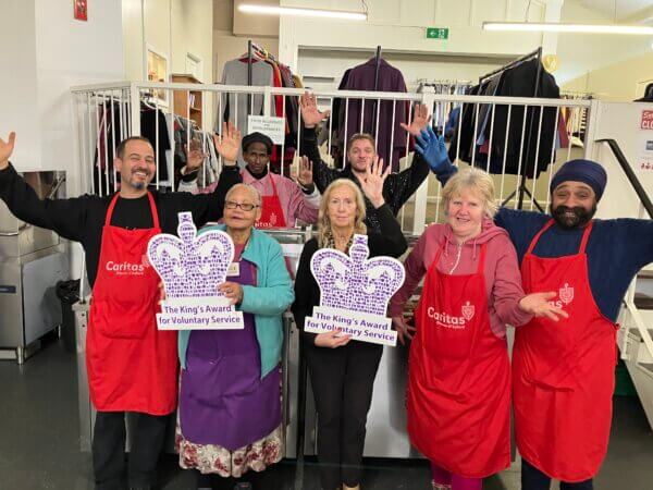 Group of people in red Caritas volunteer aprons holding Kings Award for Voluntary Service logos and smiling. They are holding their hands up in a cheering pose