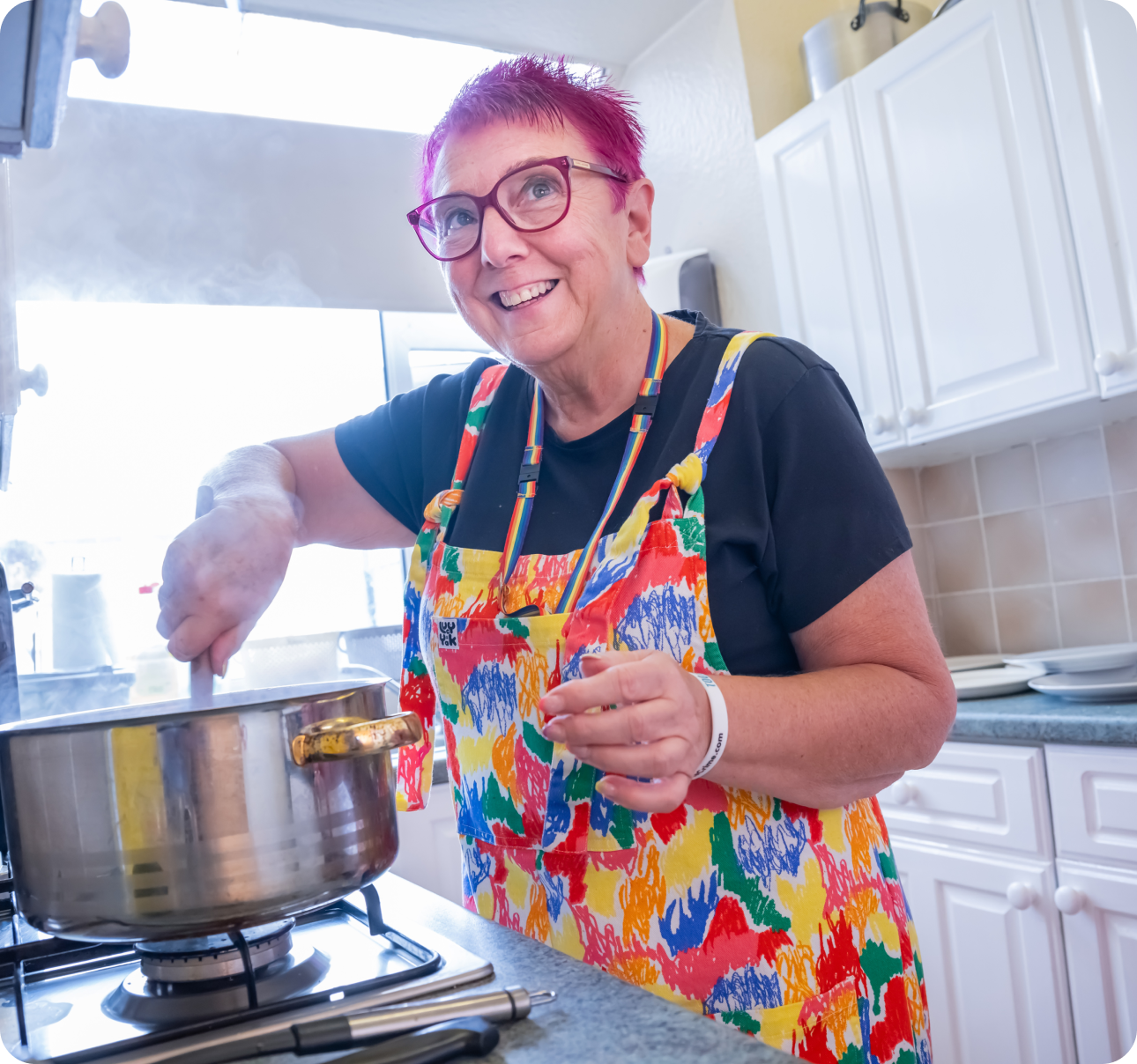 Lady with pink hair and glasses stirring a silver pan on a hob
