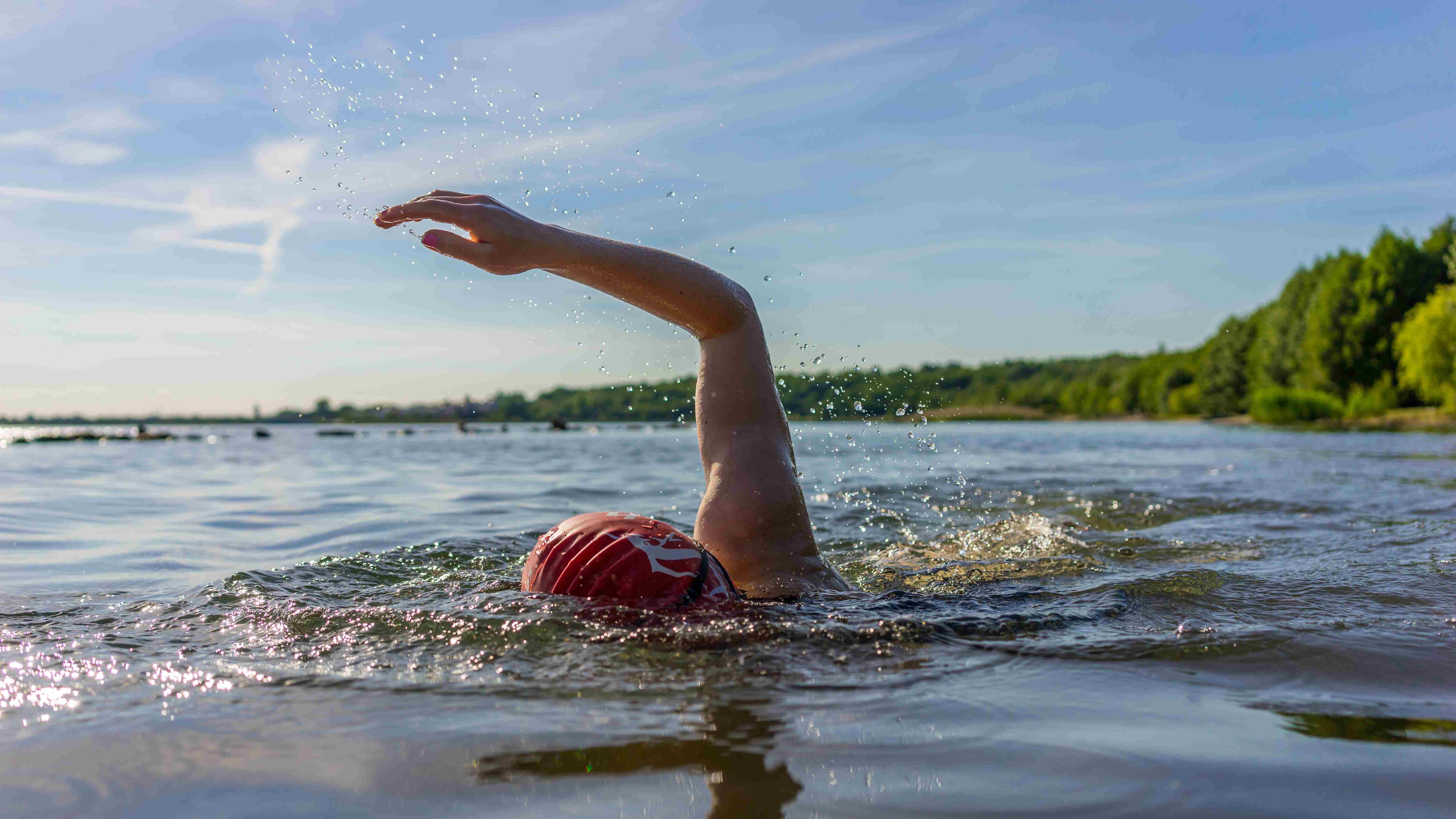 Person's head in swimming cap and arm raised as swimming a stroke. In lake with trees in the background.