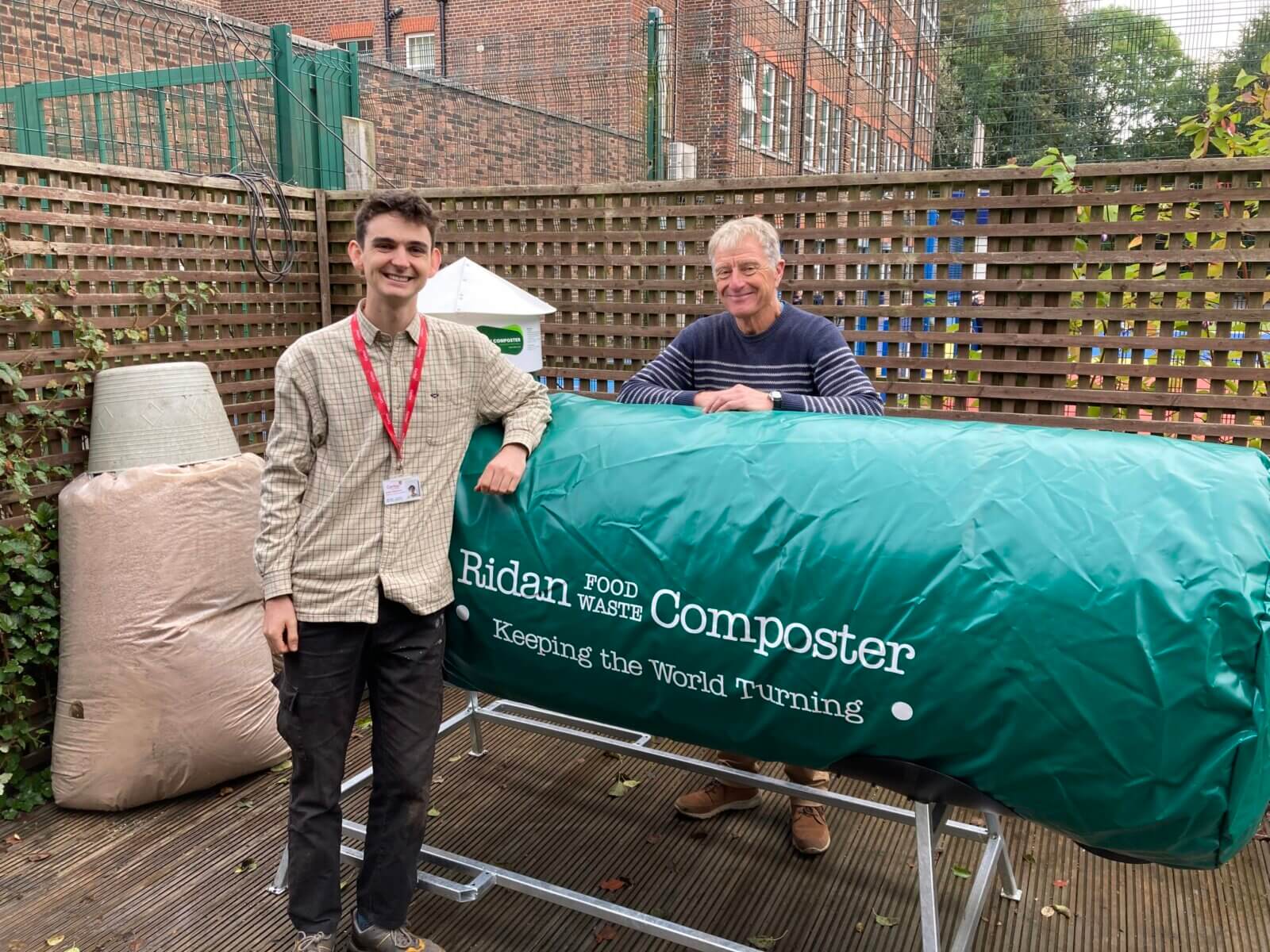 Two men standing in front of a green sleeve covered composting machine smiling at camera. The sleeve reads Ridan Food Waste Composter, keeping the world turning