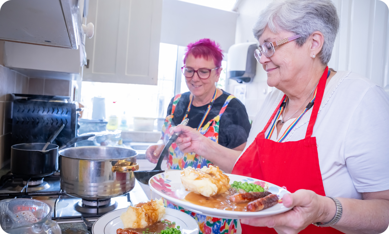 Two ladies in kitchen dishing up food. One with pink hair in a black tshirt under multicoloured top and other grey haired in red Caritas apron