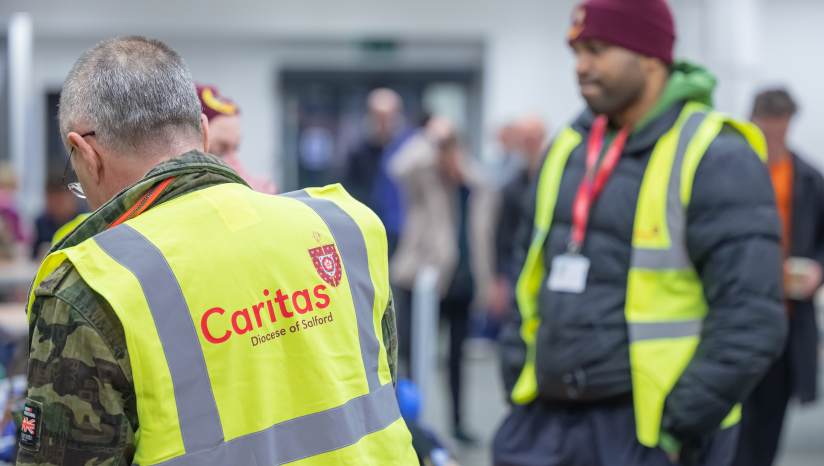 Two men in hi vis yellow jackets with caritas logo one with back to camera and one facing him