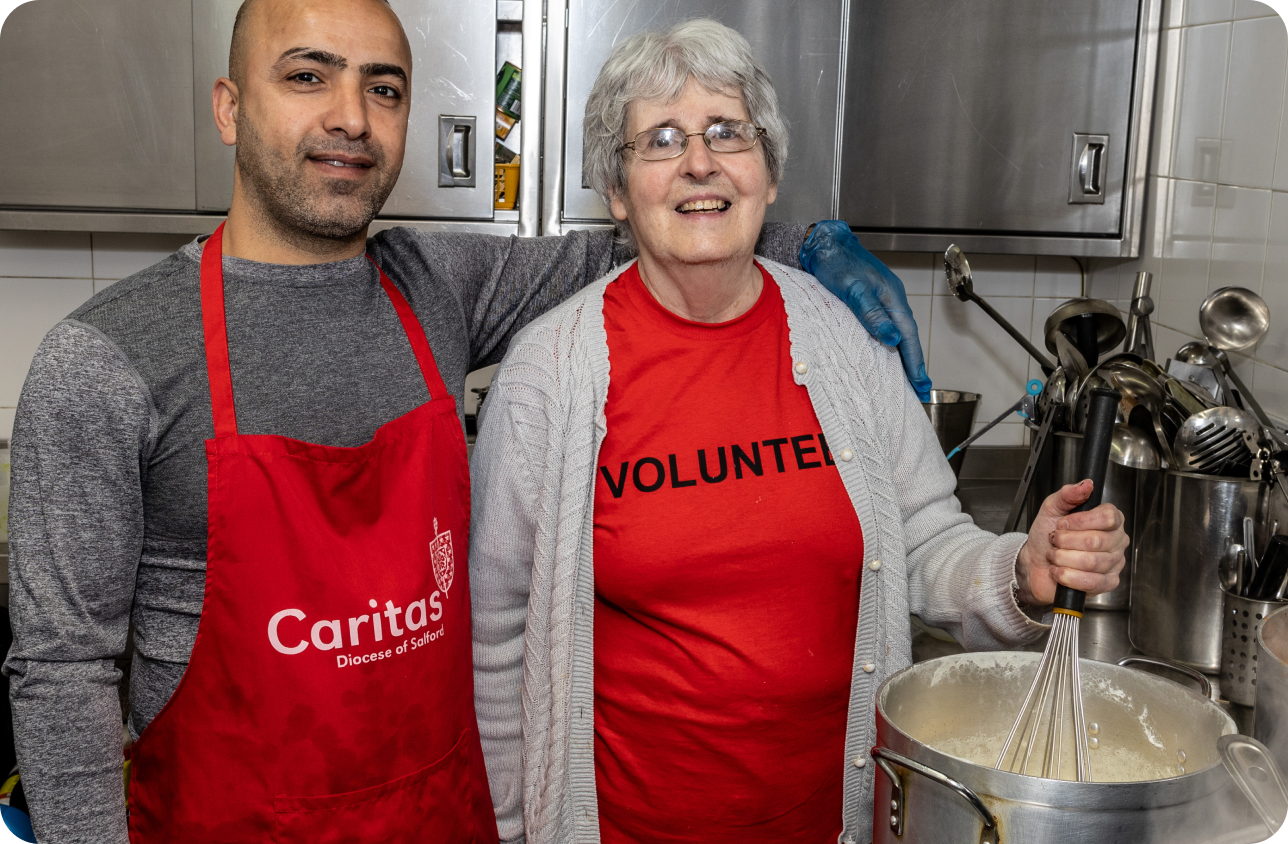 two volunteers in red caritas aprons in kitchen. One stirring metal pan
