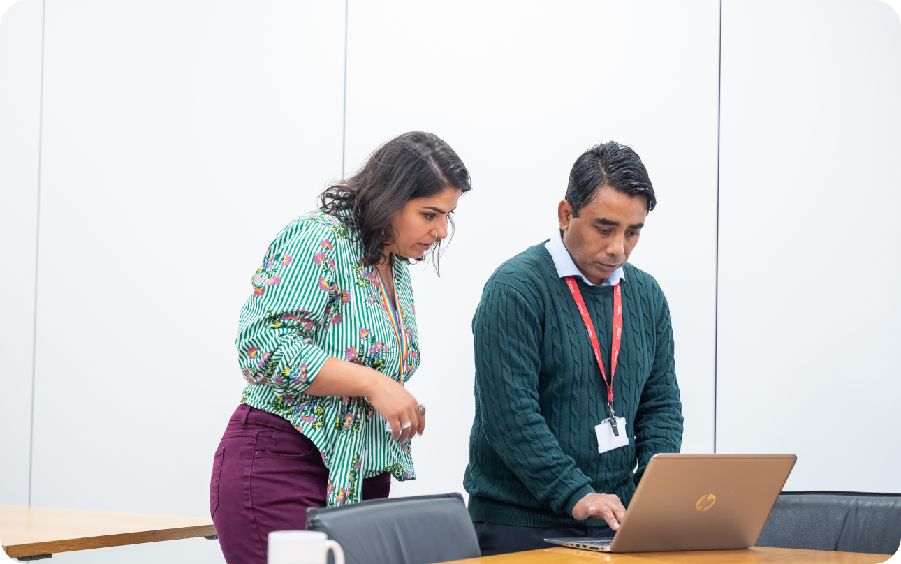 Woman in red trousers and green flowered top and man in dark grey jumper standing looking at laptop screen