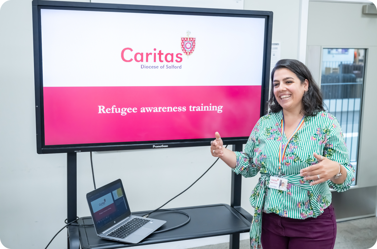 Woman in red trousers and green flowered top in front of presentation screen which says caritas and refugee awareness training