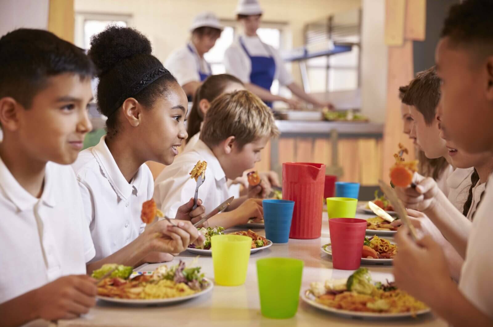 children in white polo tshirts sitting around a long table eating plates of food. Coloured drinks beakers in blue yellow red and green on the table. Blurred images of kitchen staff can be seen in the background